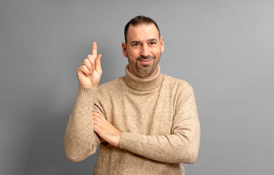 Insightful intelligent proactive man hispanic man in his 40s wearing a turtleneck sweater holding index finger up with a great new idea isolated on gray background studio portrait. People concept.