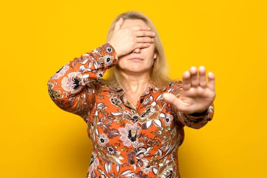 Blonde woman in a patterned shirt covering her eyes with her hands and making a stop gesture with a sad and afraid expression. Embarrassed and negative concept.