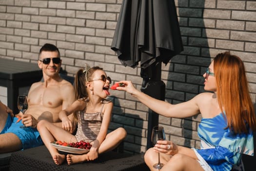 A happy family in swimsuits sunbathes on their terrace in summer. Mom feeds her daughter strawberries.