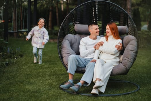 A happy family is sitting in a hammock on the lawn near the house.