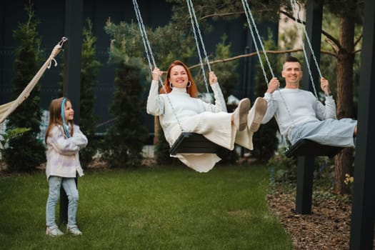 Mom and dad are riding on a swing and there is a daughter standing next to them. The family is resting on a swing.