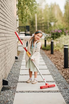 A little girl with a brush cleans a path on the street in the courtyard.