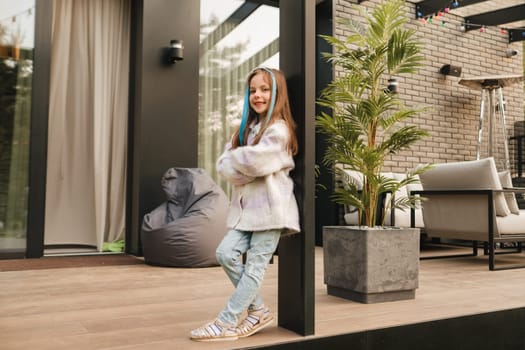 A little girl in a jacket with long hair is standing on the terrace at the house.