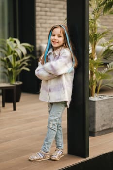 A little girl in a jacket with long hair is standing on the terrace at the house.