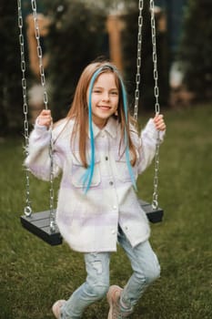 A beautiful girl with long hair in a jacket rides on a swing in the evening on the street.