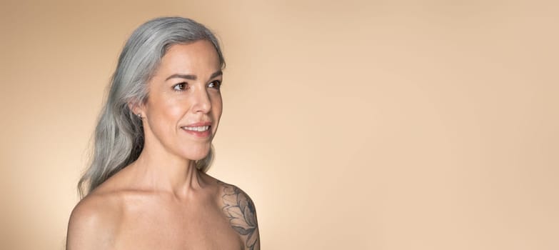 Graceful aging concept. Beautiful senior woman with glowing skin posing over beige studio background, panorama with copy space. Sensual mature woman enjoying her beauty