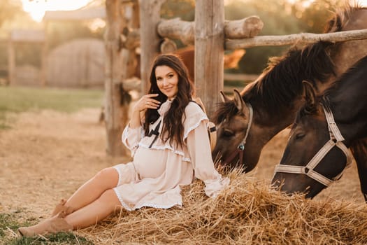 a pregnant woman in a dress in the countryside is sitting on the hay near the horses.