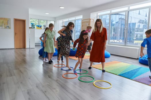 Small nursery school children with female teacher on floor indoors in classroom, doing exercise. Jumping over hula hoop circles track on the floor