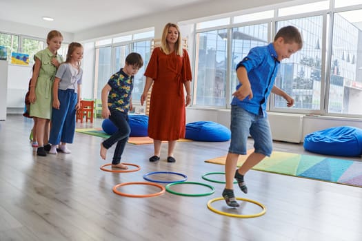 Small nursery school children with female teacher on floor indoors in classroom, doing exercise. Jumping over hula hoop circles track on the floor