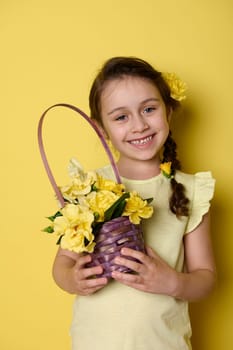 Cheerful little girl with flowers in pigtail, dressed in yellow clothes, holding purple wicker basket with yellow roses, smiling a beautiful toothy smile, looking at camera, isolated yellow background