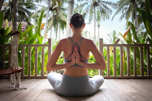 Back view of young woman doing reverse namaste mudra while meditating in tropical vacation resort. Reverse prayer pose. Yoga and spirituality concepts.