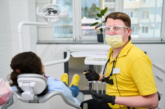 Portrait of Caucasian confident competent male dentist doctor smiling looking confidently at camera, holding dental tools, performing dental treatment to a pregnant woman sitting in dentists chair