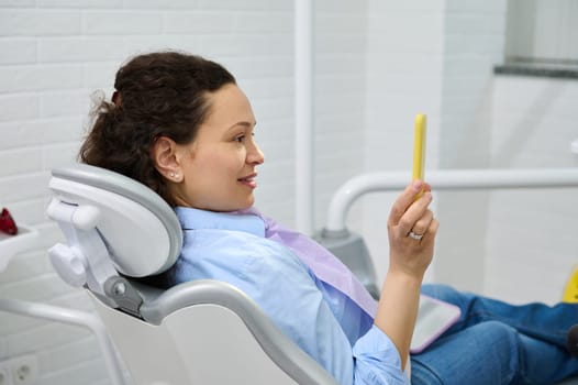 Side portrait of a happy female patient in dentist chair, smiling, looking in mirror, satisfied with treatment and professional teeth whitening in dental clinic. Oral care and dental hygiene concept