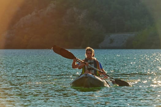 A young couple enjoying an idyllic kayak ride in the middle of a beautiful river surrounded by forest greenery in sunset time.