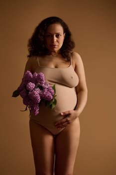Beautiful multi-ethnic body positive middle aged pregnant woman in beige underwear, holding bunch of purple blooming lilacs, touching her big belly, isolated studio background. Femininity. Pregnancy