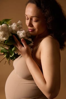 Close-up portrait of a beautiful pregnant woman in beige top, holding bunch of lilacs, posing with closed eyes over studio background. Pregnancy. Maternity. Expecting baby. Human fertility. Gynecology