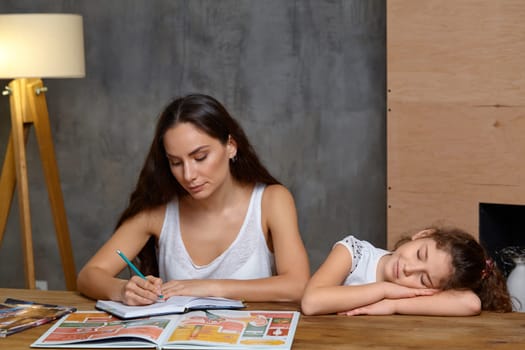 Portrait of a mother helping her small sweet and cute daughter to make her homework indoors. Mom is looking at the notebook and writing, daughter is acting like she is sleeping.