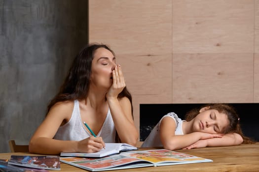 Portrait of a mother helping her small sweet and cute daughter to make her homework indoors. Mom is yawning holding a pencil in her hand, daughter is sleeping.