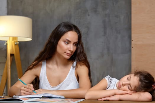 Portrait of a mother helping her small sweet and cute daughter to make her homework indoors. Mom is looking at her daughter and holding a pencil in her hand, daughter is sleeping.