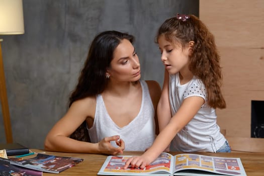 Portrait of a mother helping her small and sweet daughter to make her homework indoors. Mom is looking at her daughter and showing something in the notebook, her daughter is looking at it.