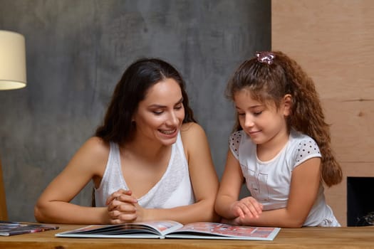 Portrait of a mother helping her small sweet and cute daughter to make her homework indoors. They are looking at the notebook and smiling.