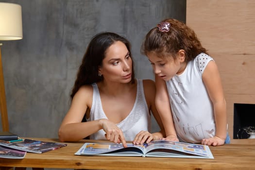 Portrait of a mother helping her small and cute daughter to make her homework indoors. Mom is showing something in the notebook and her daughter is looking at it.