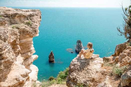 Happy girl perched atop a high rock above the sea, wearing a yellow jumpsuit and braided hair, signifying the concept of summer vacation at the beach