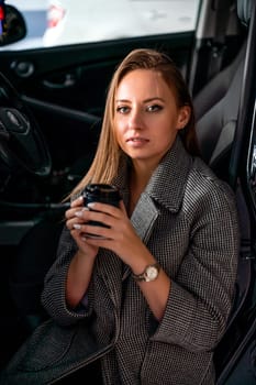 Happy woman coffee. she stands next to the car in the underground parking. Dressed in a gray coat, holding a glass of coffee in her hands, a black car