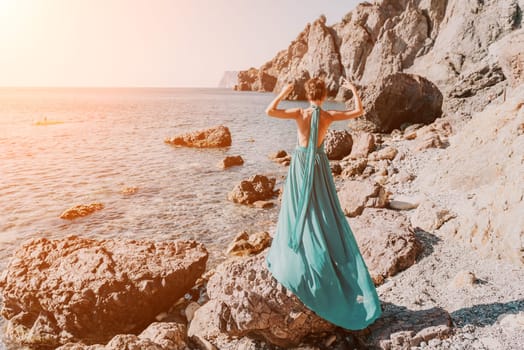 Side view a Young beautiful sensual woman in a red long dress posing on a rock high above the sea during sunrise. Girl on the nature on blue sky background. Fashion photo.