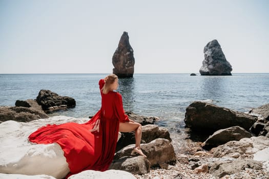 Woman travel sea. Young Happy woman in a long red dress posing on a beach near the sea on background of volcanic rocks, like in Iceland, sharing travel adventure journey