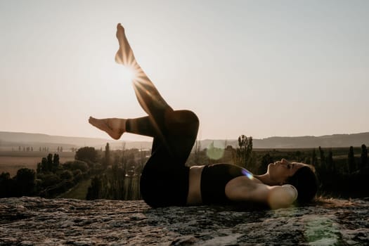 Well looking middle aged woman with long hair, fitness instructor in leggings and tops doing stretching and pilates on the rock near forest. Female fitness yoga routine concept. Healthy lifestyle.