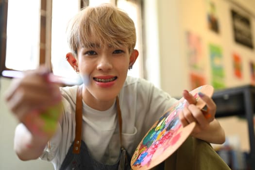 Cheerful teenage gay man holding paintbrushes and palette. Education, hobby, art concept.