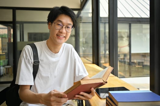 Smiling asian man student in glasses sitting at coffee shop and reading book. Education, learning and technology concept.