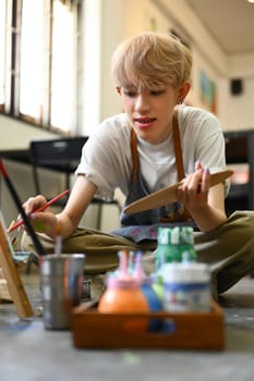 Image of young male artist holding palette and painting picture on canvas with oil paints in art studio.