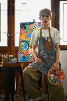 Portrait of young hipster queer artist wearing apron holding palette and brush posing in front of easel in art studio.