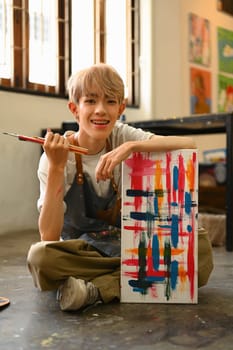 Full length view of young gay male artist sitting on the floor in art studio lit by sunlight.