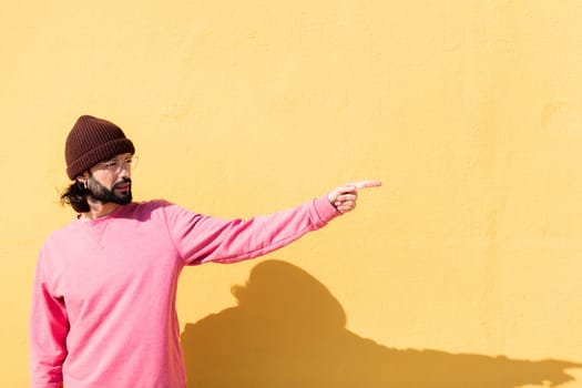 stylish young man with pink sweater pointing to the side in a yellow background, concept of youth and modern lifestyle, copy space for text
