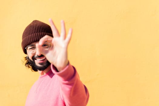 stylish young man with pink sweater doing ok sign with hand in a yellow background, concept of youth and modern lifestyle, copy space for text