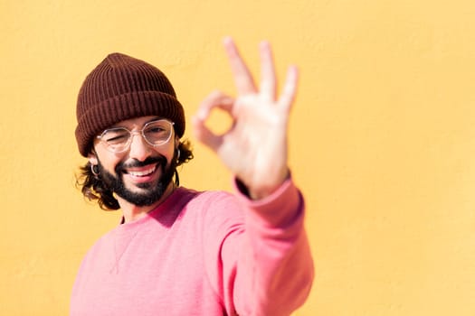 stylish young man with pink sweater doing ok sign with hand in a yellow background, concept of youth and modern lifestyle, copy space for text