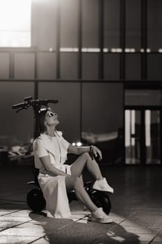 A stylish girl in glasses and a white summer coat in the city is sitting on an electric scooter.