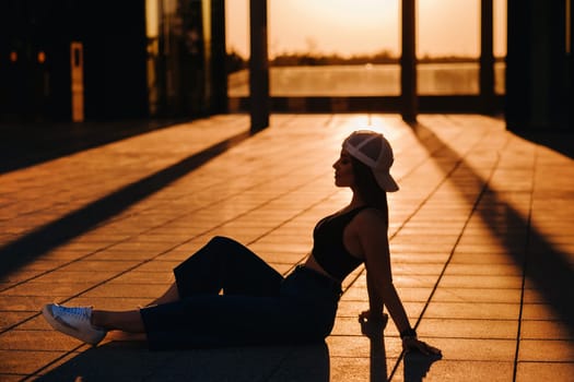 The silhouette of a young teenage girl in jeans and a cap is sitting on the asphalt at sunset.