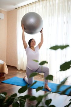 View through a houseplant on the foreground, to a pregnant woman, expectant mother, smiling while exercising with a fitball at home. Gravid mom enjoying prenatal fitness and active maternity lifestyle