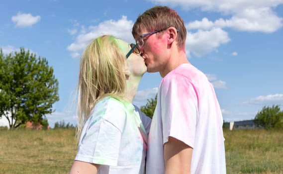 Romantic Caucasian Couple Adults Kiss With Colorful Paint, Powder On Clothes On Holi Color Festival. Emotional Happy Man and Woman. Blue Sky, Green Trees On Background, Sunny Day. Horizontal Plane.