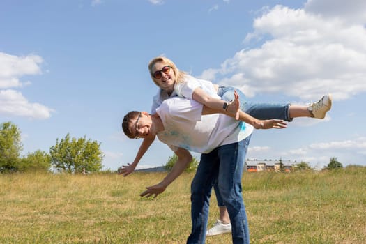 Happy Man Holds Woman On His Back With Colorful Dye, Powder On Clothes On Holi Colors Festival. Cheerful Couple. Blue Sky, Green Trees On Background, Summertime. Copy Space For Text. Horizontal Plane.