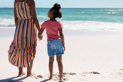 African american girl holding hand of mother while standing together at beach on sunny day. unaltered, family, lifestyle, togetherness, enjoyment and holiday concept.