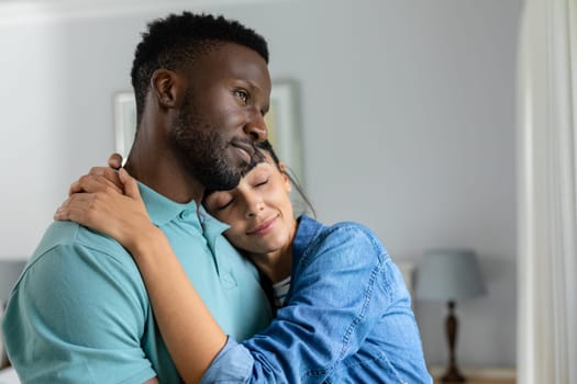 Thoughtful multiracial young couple embracing while standing in living room at home. unaltered, lifestyle, domestic life, togetherness, love, contemplation.