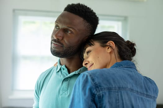 Multiracial young couple looking away while embracing in living room at home. unaltered, lifestyle, domestic life, togetherness, love, contemplation.
