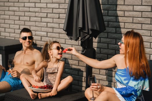 A happy family in swimsuits sunbathes on their terrace in summer. Mom feeds her daughter strawberries.