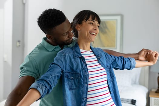 Happy multiracial young couple with arms outstretched standing in living room at home. unaltered, lifestyle, domestic life, togetherness, love.