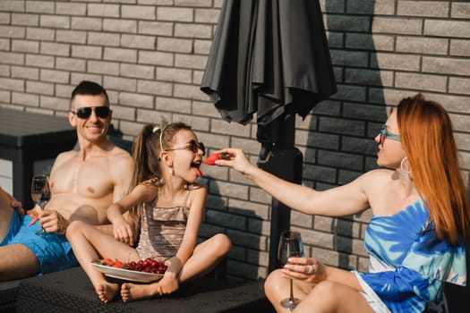 A happy family in swimsuits sunbathes on their terrace in summer. Mom feeds her daughter strawberries.